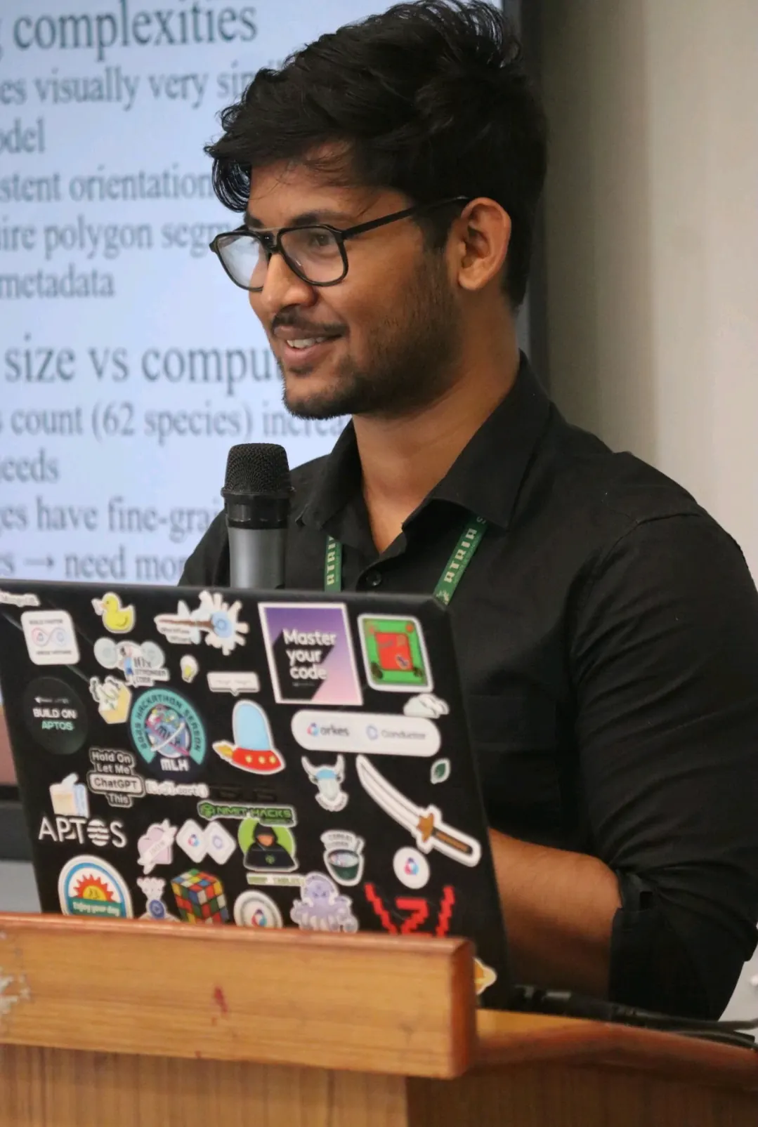 Ujesh speaking into a microphone, his sticker-covered laptop beside him